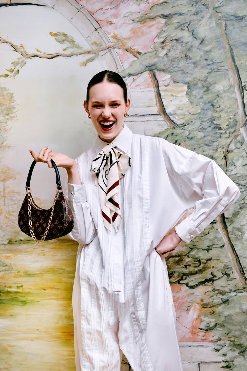 Woman in white jumpsuit poses with patterned handbag against a painterly backdrop, smiling with open mouth in a studio setting.