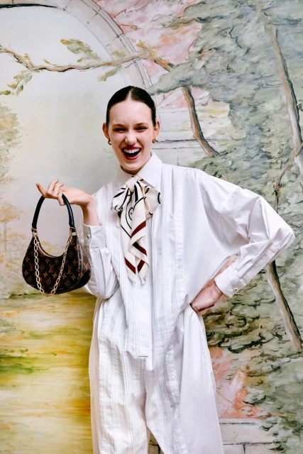 Woman in white jumpsuit poses with patterned handbag against a painterly backdrop, smiling with open mouth in a studio setting.