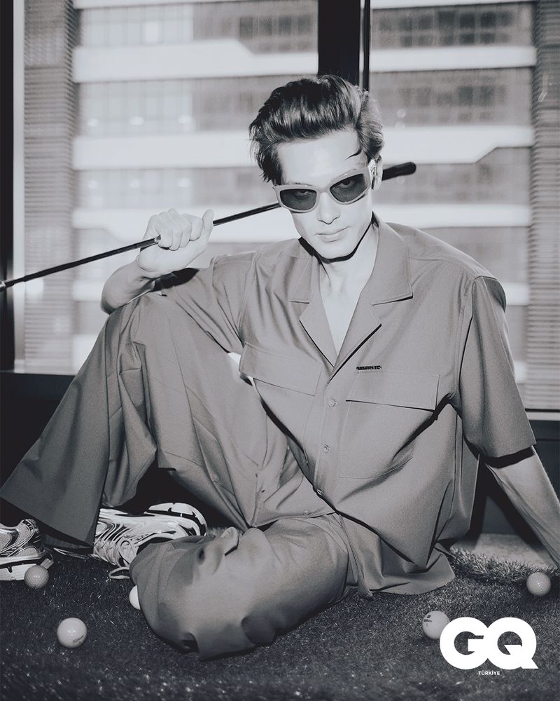 Black and white portrait of a man with sunglasses, casually dressed, seated indoors near a window with a golf club and balls.