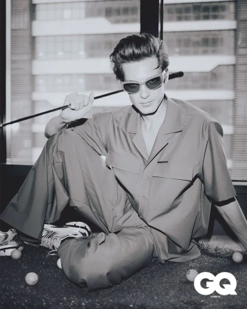 Black and white portrait of a man with sunglasses, casually dressed, seated indoors near a window with a golf club and balls.
