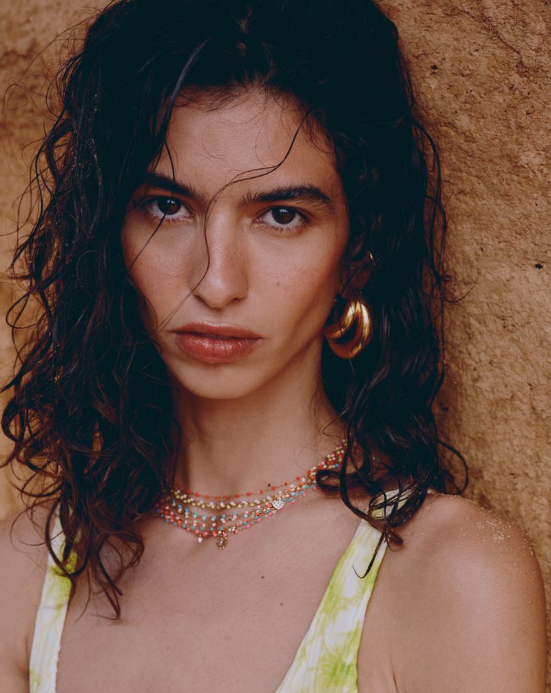 Close-up portrait of a young brunette woman with gold earrings and a beaded necklace, posing against a tan wall in soft, warm light.