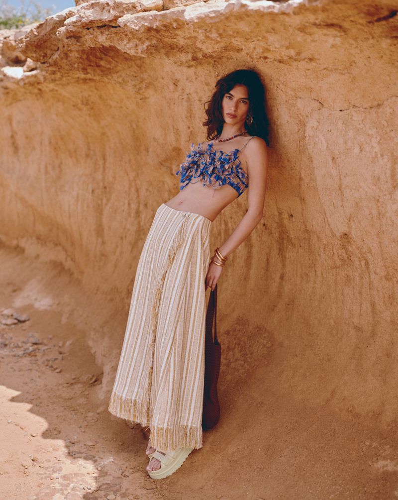 A woman poses against a rock formation, wearing a blue floral top and striped skirt, complemented by brown and beige desert tones.