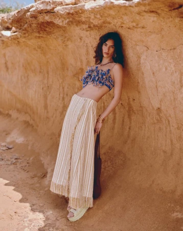 A woman poses against a rock formation, wearing a blue floral top and striped skirt, complemented by brown and beige desert tones.
