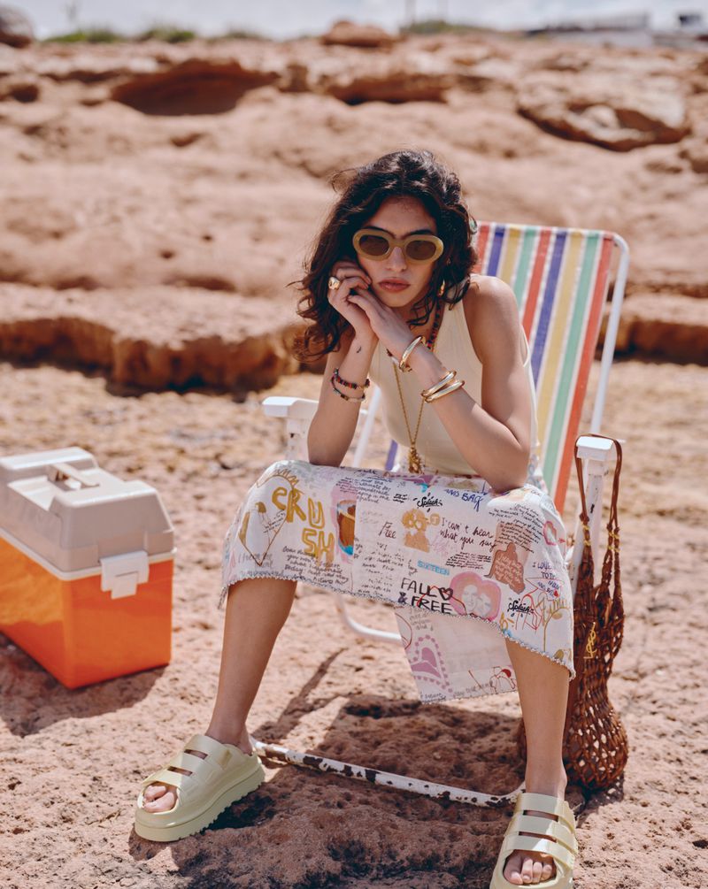Woman in sunglasses and skirt with graphic print sits on striped beach chair on sandy beach next to a cooler on a sunny day.