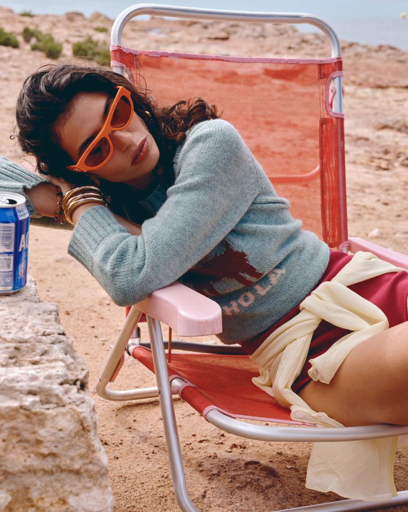 Woman in sunglasses and a light blue sweater with 'Hola' on it, relaxing in a red folding beach chair on a sandy beach.