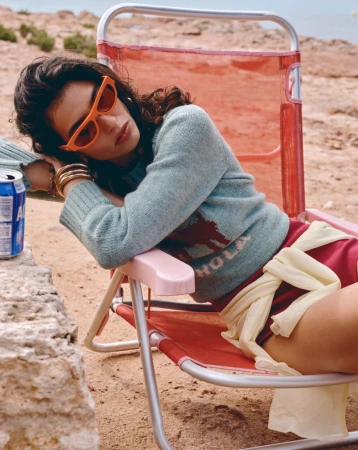 Woman in sunglasses and a light blue sweater with 'Hola' on it, relaxing in a red folding beach chair on a sandy beach.