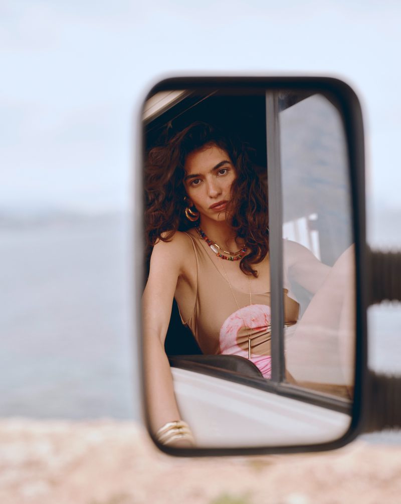 Woman reflects in a white vehicle mirror with wavy brown hair, wearing a tan top, a beaded necklace, and gold earrings.