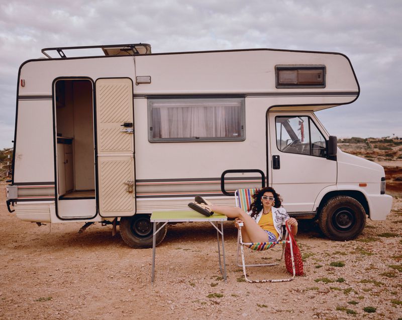 Woman sits outside retro camper van in shorts, patterned shirt, and sunglasses, relaxing in a chair with a red bag.