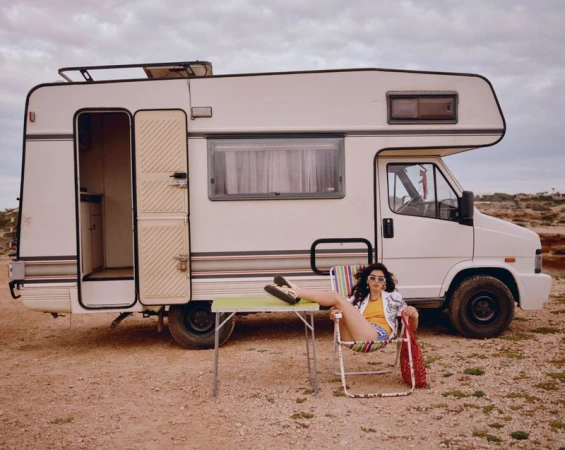 Woman sits outside retro camper van in shorts, patterned shirt, and sunglasses, relaxing in a chair with a red bag.