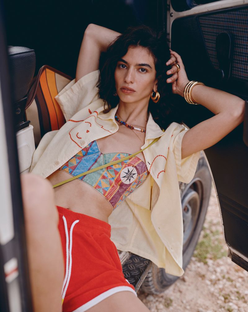 A stylish young woman in a colorful top and red shorts reclines against a vehicle, showcasing a blend of fashion and casual charm.