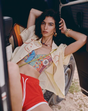 A stylish young woman in a colorful top and red shorts reclines against a vehicle, showcasing a blend of fashion and casual charm.