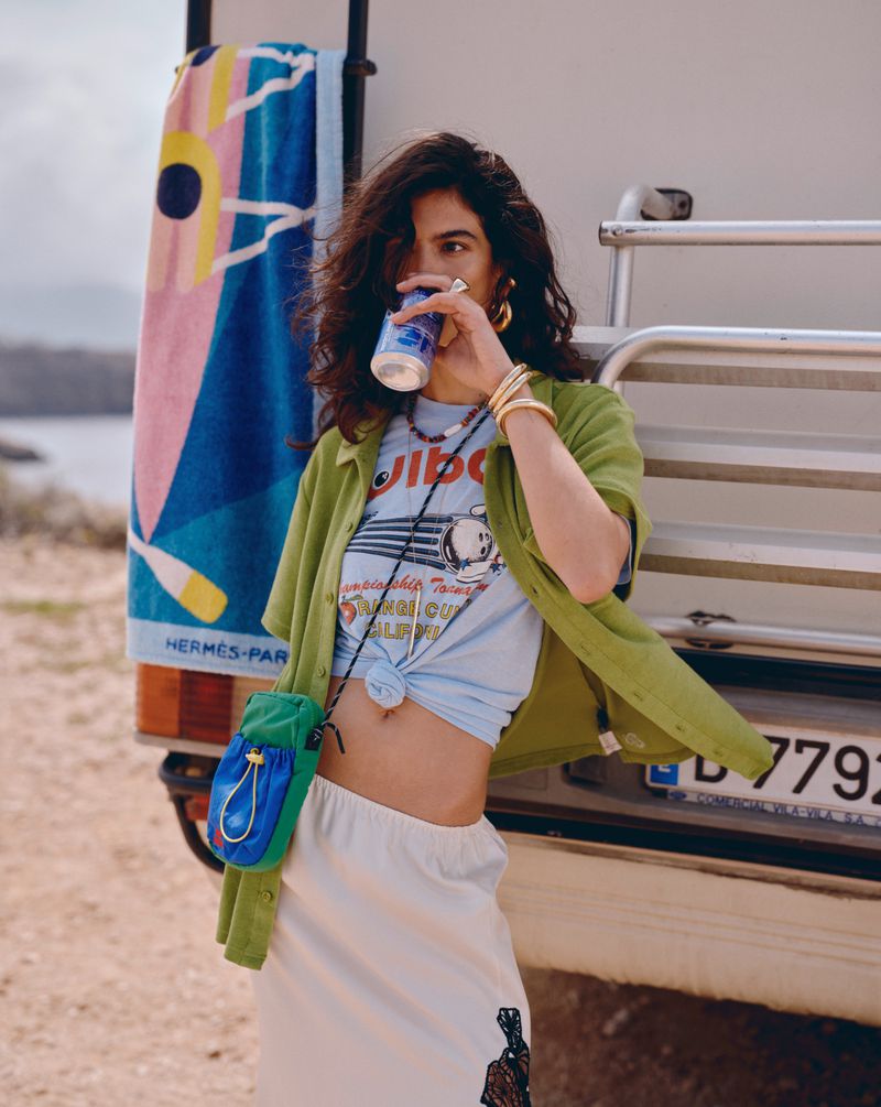 Brunette woman drinking from a can near a camper with a beach towel hanging on the back, wearing a skirt and casual clothing.
