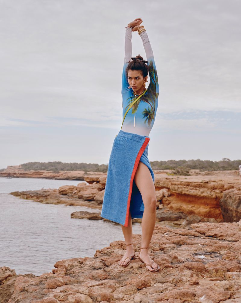 Adult woman posing on a rocky beach with ocean and sky background, wearing a long-sleeve top and a blue sarong-style skirt.