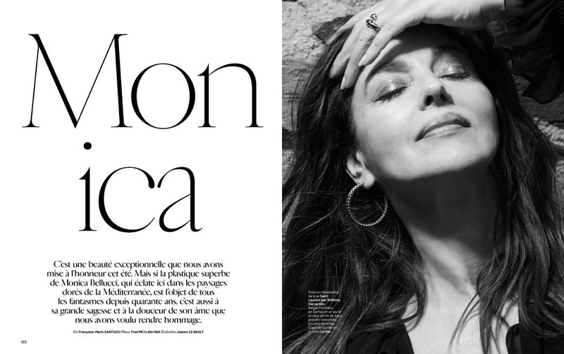 Black and white portrait of Monica Bellucci with closed eyes, hand on forehead, hoop earrings, against a textured stone background.