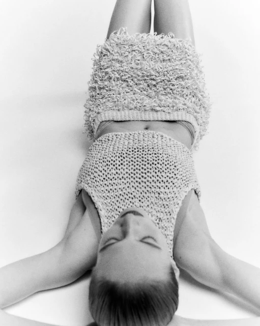 Black and white portrait of a woman lying down wearing a knitted crop top and textured mini skirt, eye closed, neutral expression.