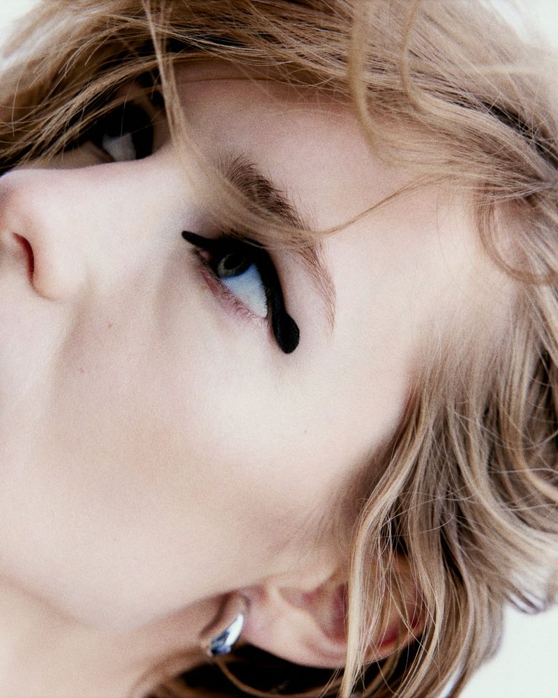 Close-up shot of a blonde woman's face, featuring a dark eyeliner design that accentuates her eye, alongside a silver earring.