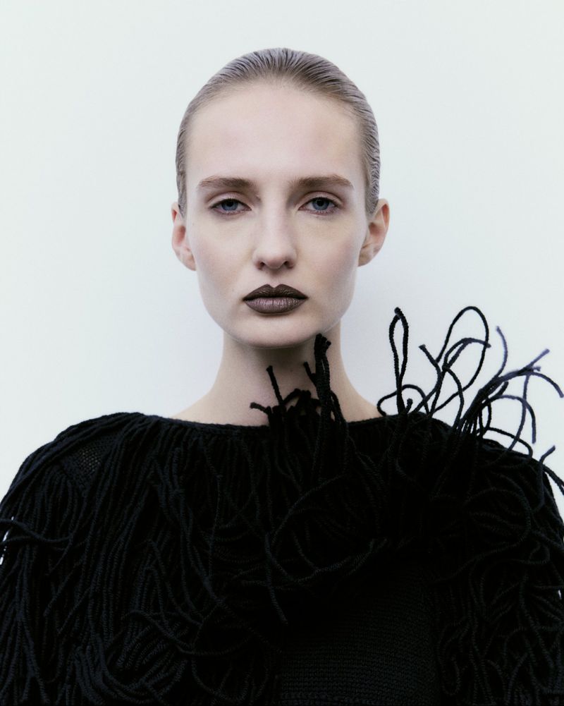 Close-up portrait of a fashion model with dark lipstick wearing a textured black fringe sweater against a white background.