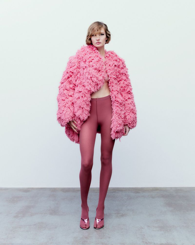 A fashion photo of a woman in a pink faux fur coat, pink leggings, and pink high-heeled sandals, posing in front of a neutral background.