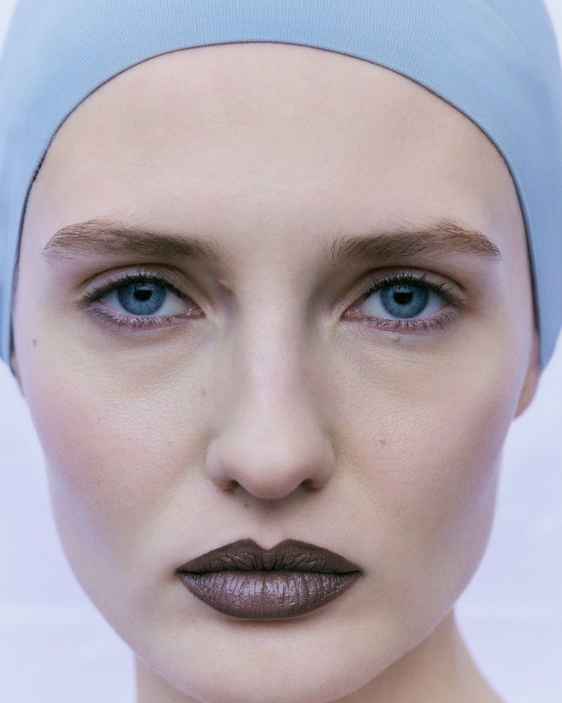 Close-up portrait of a woman with light skin, blue eyes, and dark lipstick, wearing a light blue head covering against a light background.