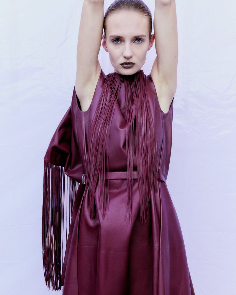 A woman with pale skin and dark lipstick poses in a studio wearing a sleeveless dress with fringe on the bodice.