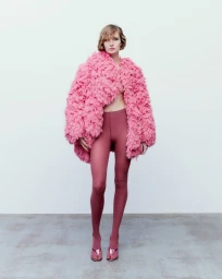 Model Roe Elema wearing an oversized textured pink shaggy coat and dusty rose tights in a minimalist studio setting.