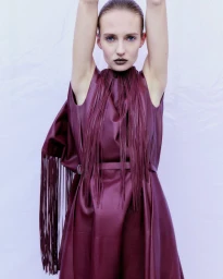 Model Roe Elema poses with arms raised wearing a burgundy leather dress with long fringe detailing against a pale studio backdrop.