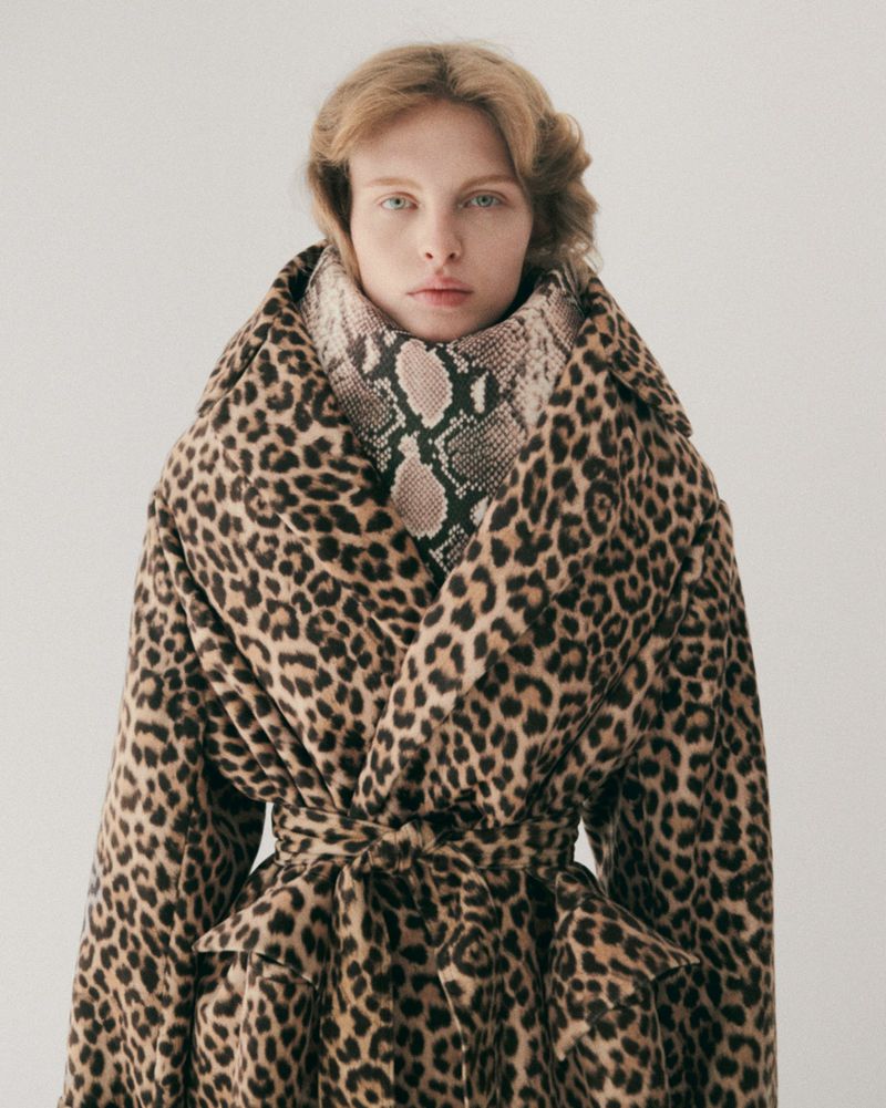 Woman wearing an animal print coat and snakeskin scarf stands against a white background, looking at the camera in a studio shot.