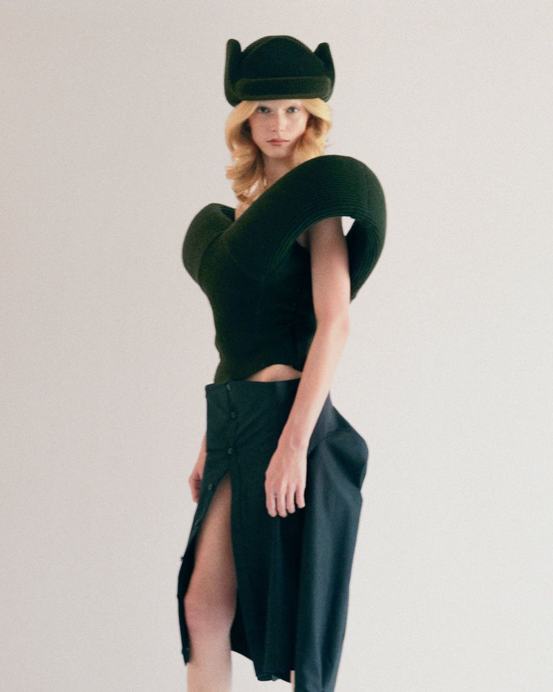 A woman with blonde hair poses in a dark green sculptural outfit consisting of a sleeveless top, midi skirt with buttons, and a hat.