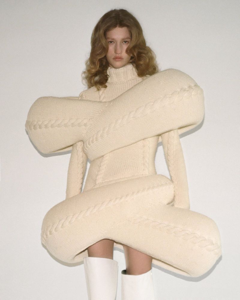 Model showcasing oversized knit dress in studio. Woman in textured ivory dress, white boots, and soft waves poses against a white background.