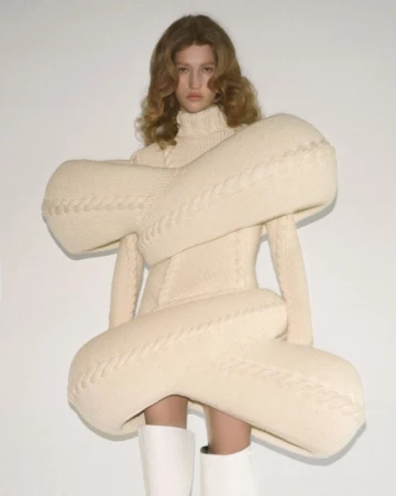 Model showcasing oversized knit dress in studio. Woman in textured ivory dress, white boots, and soft waves poses against a white background.