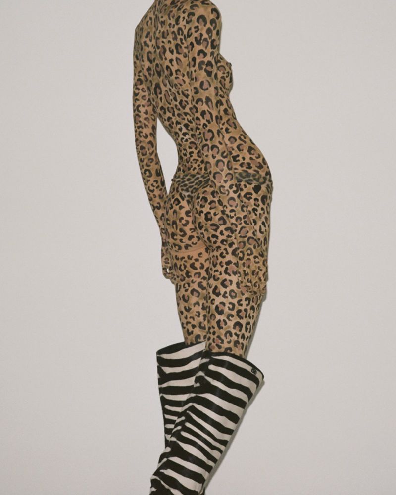 Fashion model wearing a tan animal print bodysuit and black and white zebra print boots posing against a light neutral background.