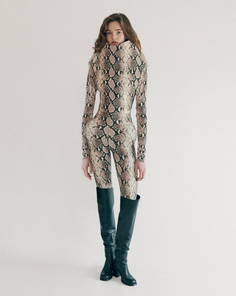 A full-length portrait of a slender young woman with long, wavy hair, wearing a snakeskin-patterned jumpsuit and black, knee-high leather boots.