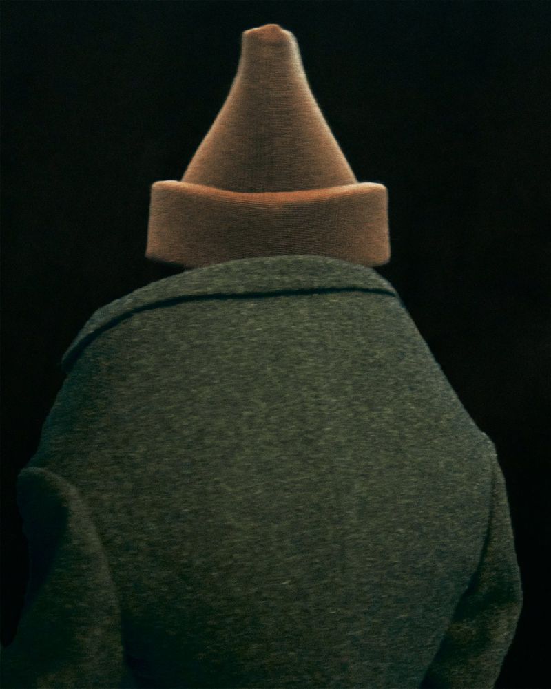 Back view of a person wearing a grey coat and a brown dunce cap against a dark background. The hat is cone-shaped and sits squarely.