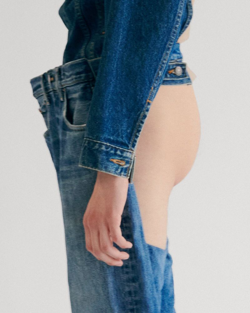 A person wearing a denim jacket and jeans with an unusual cut-out design, posing against a bright white backdrop in a studio setting.