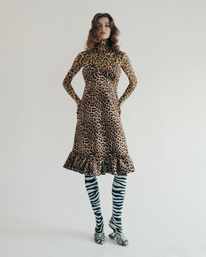 A woman in a patterned long-sleeve top and ruffled midi dress stands in front of a blank white background, wearing zebra-striped tights and heels.
