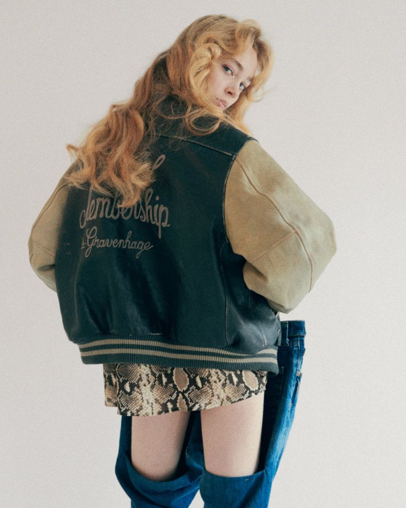 Blonde woman in a varsity jacket and animal print skirt poses in a studio against a white background, looking over her shoulder.