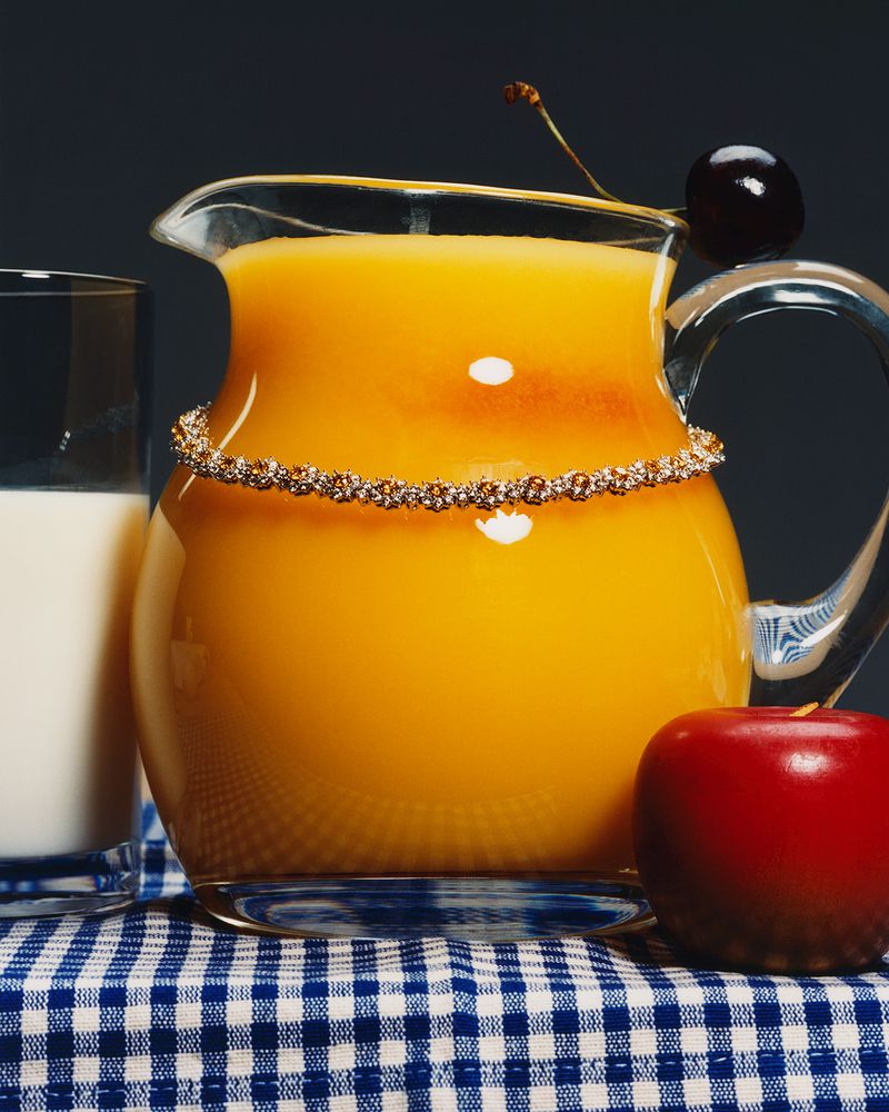 Close-up still life with a glass of milk, a pitcher of orange juice garnished with a cherry, and an apple on a checkered tablecloth.