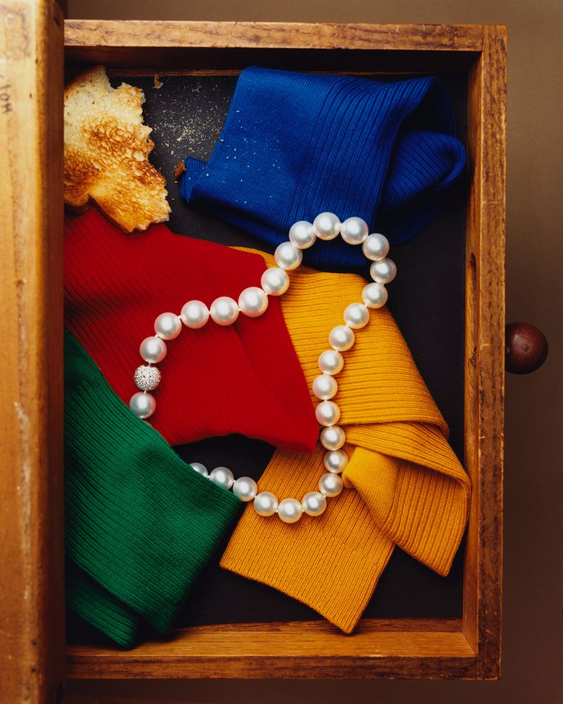 Overhead shot of a pearl necklace draped over colorful folded knitwear inside of an open wooden drawer with bread in the corner.