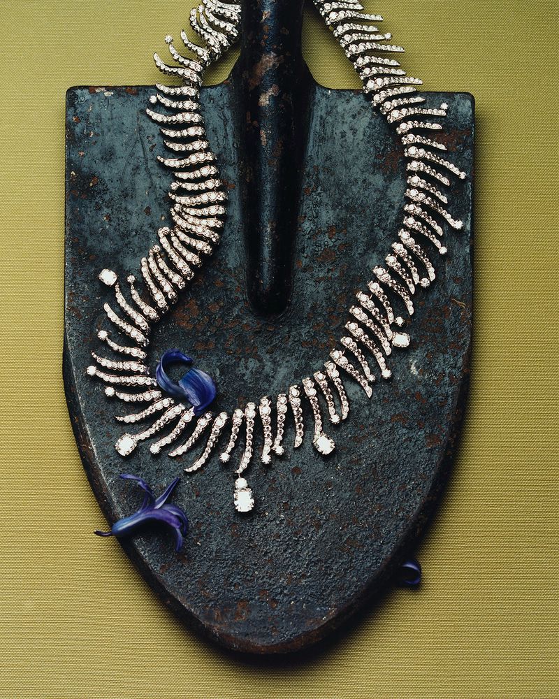 Elegant diamond necklace draped on a weathered shovel, accented with small blue flowers, set against a textured green backdrop.