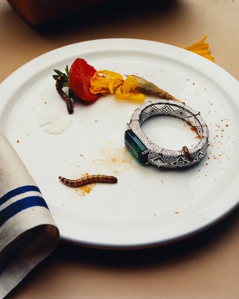 Elevated close-up of an ornate diamond bracelet, strawberries, yellow flower petals, and a mealworm on a white plate with a napkin.