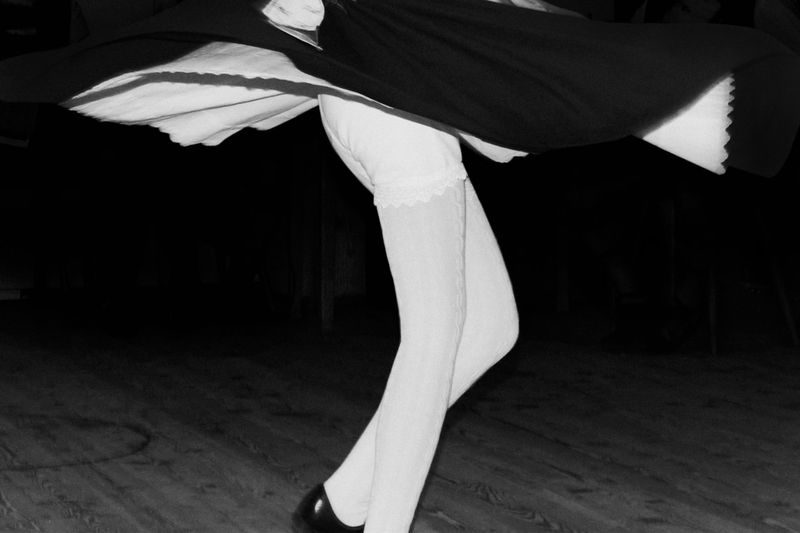 A woman wearing a dark dress and white stockings twirls on a wooden floor in this high-contrast black and white studio photograph.