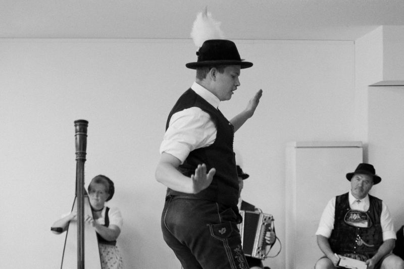 Black and white photo of a man in traditional Bavarian clothing dancing, accompanied by musicians with a harp and accordion.