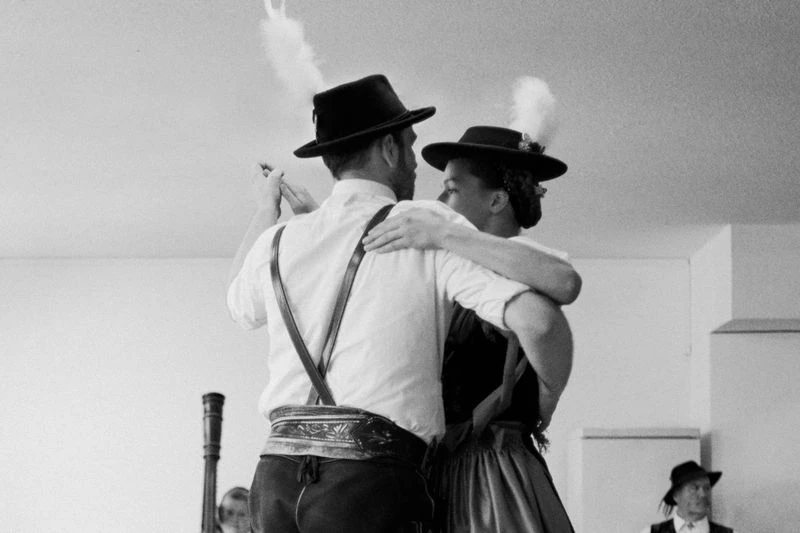 A couple wearing traditional German Tracht clothing dances in a bright indoor space, another individual visible in the background.