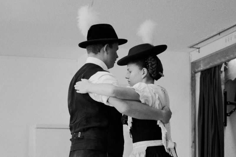 A couple dressed in traditional attire embraces indoors, showcasing vintage clothing and hats with feather accents in a monochrome image.