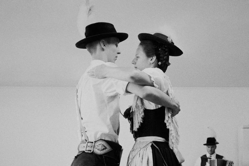 A black and white shot of a couple dancing, dressed in folk costumes, indoors with a musician visible in the background.