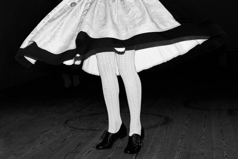 Monochrome image of a person wearing a vintage dress, patterned tights, and black shoes, standing on a wooden floor in a dark room.