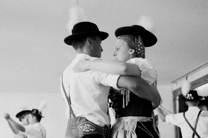 Monochromatic image of a man and woman in traditional German attire, embracing indoors; a dance scene with soft lighting and period costumes.
