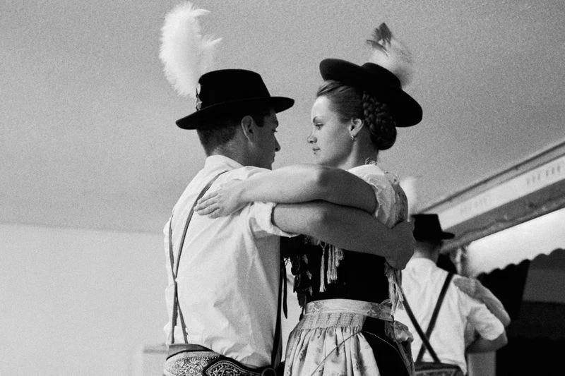Couple in traditional German attire embracing, wearing feathered hats, indoors in a vintage-style black-and-white photo, folk dancing.