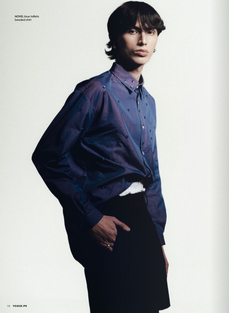 Masculine-presenting fashion model posing in a blue button-down shirt and black pants against a clean white backdrop in a studio setting.