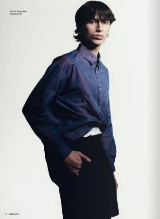 Masculine-presenting fashion model posing in a blue button-down shirt and black pants against a clean white backdrop in a studio setting.